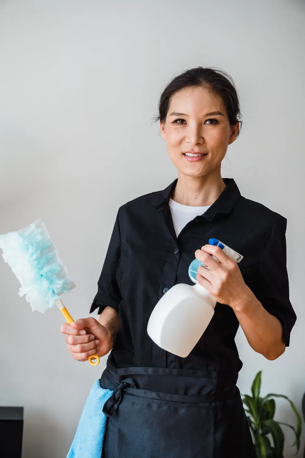 Smiling cleaner in a black uniform and apron holding a duster and a spray bottle